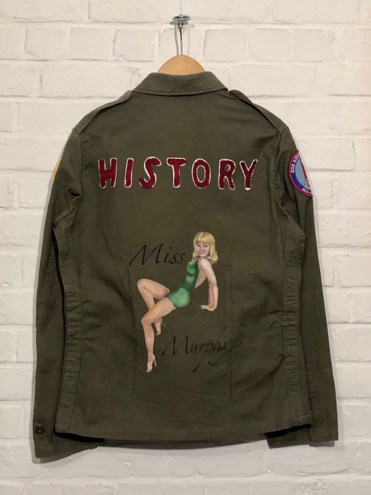 Green jacket with a patch on a hanger against a white brick wall.
