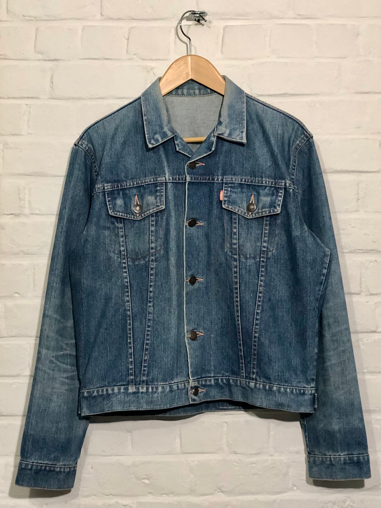 Blue denim jacket on a hanger against a light brick wall.